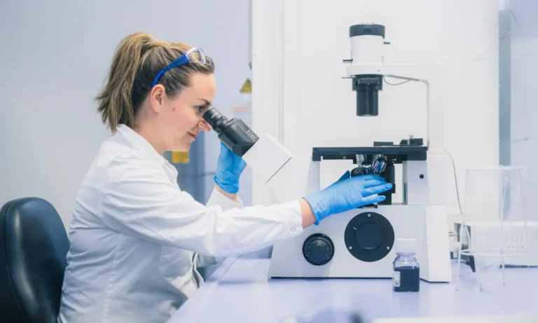 Ein Wissenschaftler in weißem Laborkittel, blauen Handschuhen und Schutzbrille untersucht in einem Auftragslabor eine Probe unter dem Mikroskop.