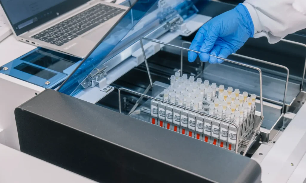 Eine behandschuhte Hand hält ein mit Reagenzgläsern gefülltes Metallgestell in einem Analysegerät eines Auftragsanalytik-Labors, daneben steht ein Laptop, und eine durchsichtige blaue Abdeckung ist teilweise geöffnet.