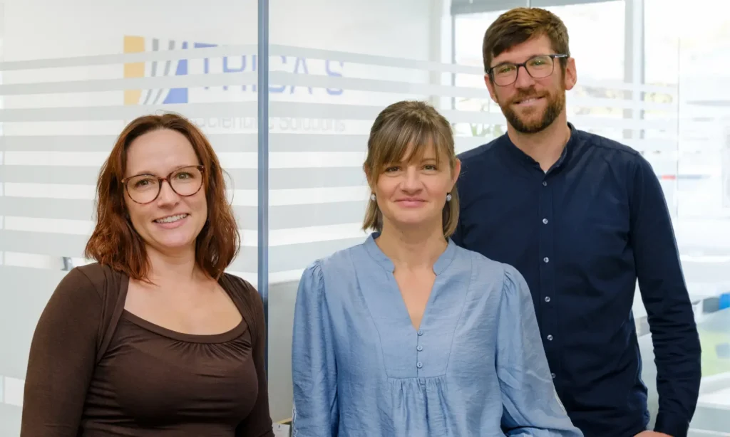 Drei Personen – zwei Frauen und ein Mann – stehen in einem Innenraum vor einer gläsernen Bürowand und lächeln in die Kamera. Alle tragen eine Brille und Business-Casual-Kleidung, was die professionelle Atmosphäre eines Biostatistik-Teams widerspiegelt. Auf der Glaswand ist das Firmenlogo zu sehen.