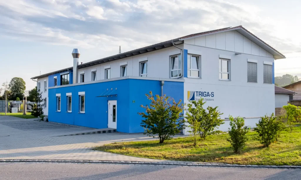 Ein modernes zweistöckiges Gebäude mit weißen und blauen Außenwänden, das mit „TRIGAS FI Flow Solutions“ beschriftet ist und möglicherweise modernste TRIGAS-IVD-Technologie beherbergt, steht inmitten von Gras, Sträuchern und einem gepflasterten Gehweg unter einem teilweise bewölkten Himmel.