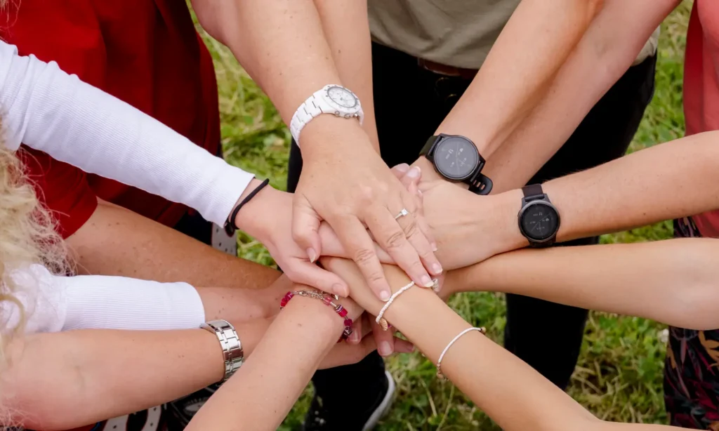 Eine Gruppe von Menschen, die im Kreis steht, legt ihre Hände in der Mitte übereinander, was die Teamarbeit und den Zusammenhalt von „Karriere Triga“ symbolisiert. Man sieht verschiedene Uhren, Armbänder und Hautfarben, darunter ist Gras zu sehen.
