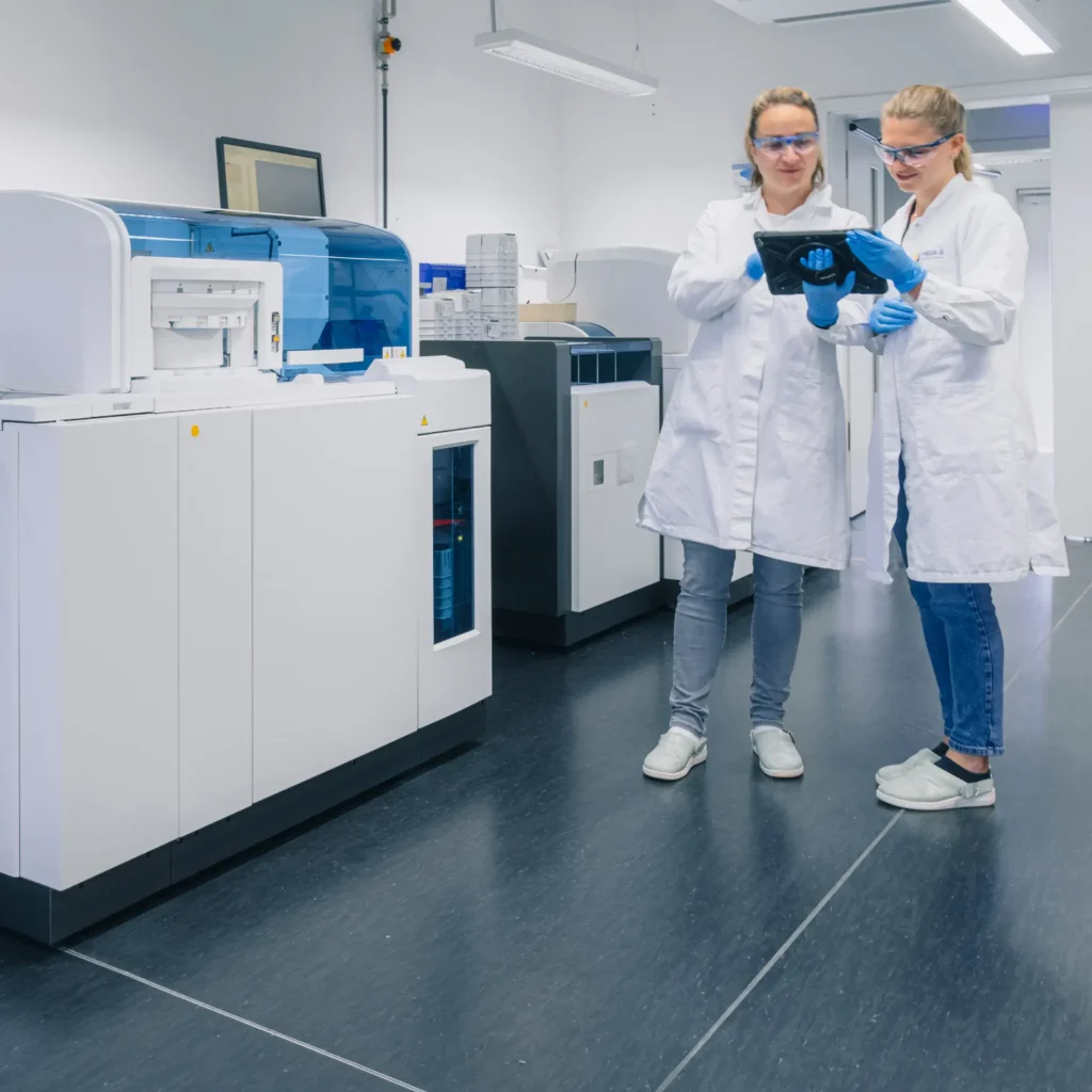 Zwei Wissenschaftler in Laborkitteln, Handschuhen und Schutzbrillen stehen in einem modernen Labor, schauen gemeinsam neben großen Analysegeräten auf ein Tablet und besprechen Daten für IVDR-Leistungsstudien.