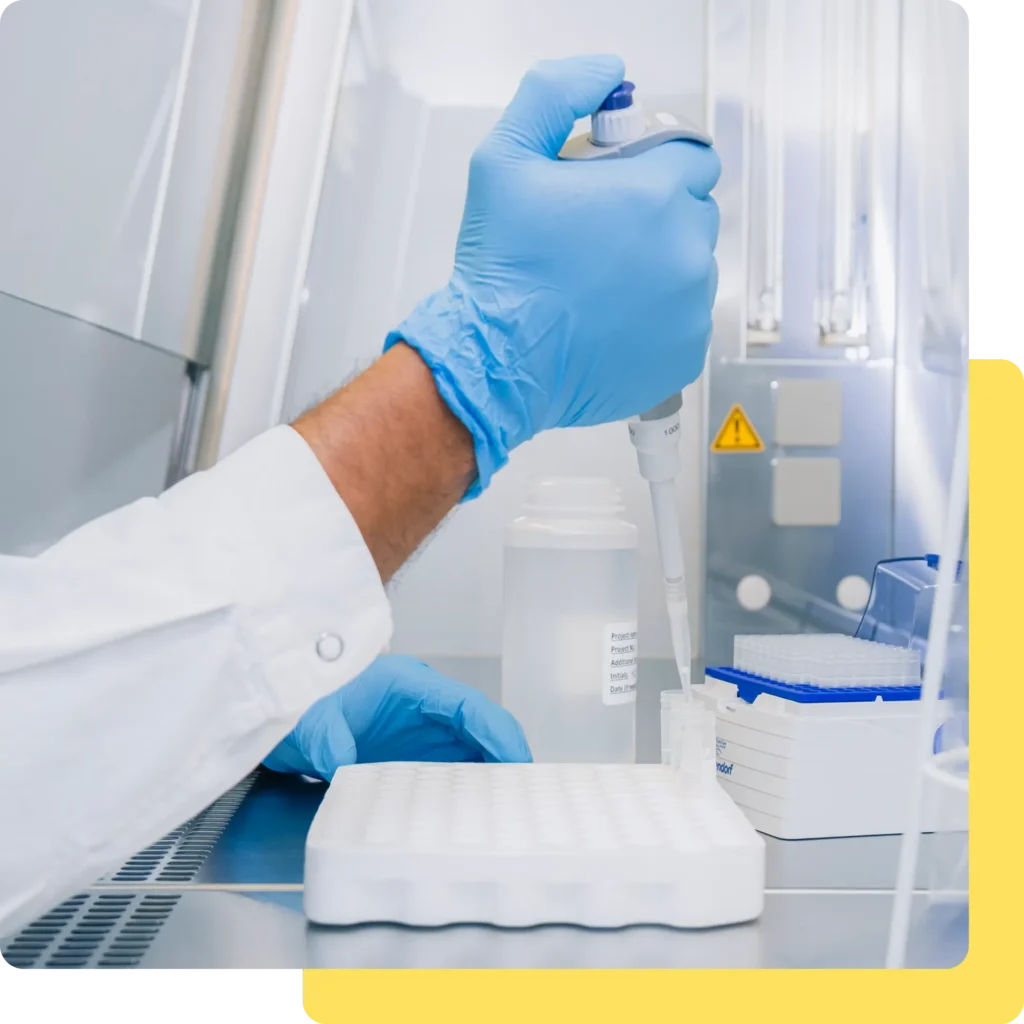 Eine Person in einem Laborkittel und blauen Handschuhen füllt mit einer Pipette Flüssigkeit in eine Platte mit mehreren Vertiefungen in einer Laborumgebung und führt dabei IVDR-Leistungsstudien durch, umgeben von Laborgeräten und Sicherheitsschildern.