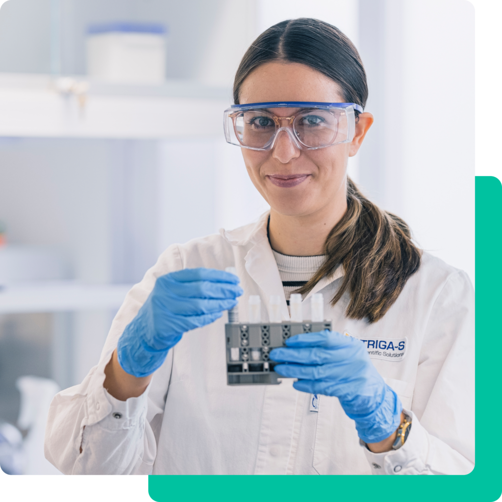 Eine Frau in einem Laborkittel, mit Schutzbrille und blauen Handschuhen hält in einem hellen Laborraum ein Reagenzglasgestell mit Triga-S-Laborgeräten in der Hand.