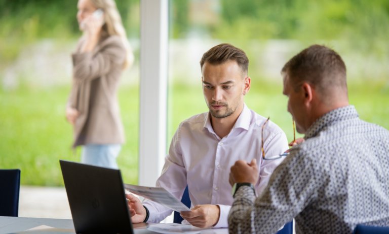 Zwei Männer sitzen drinnen an einem Tisch und konzentrieren sich auf Unterlagen und einen Laptop mit DMID-Daten. Der eine hält Papiere in der Hand, während der andere mit seiner Brille gestikuliert. Draußen steht eine Frau und telefoniert, im Hintergrund ist Grün zu sehen.