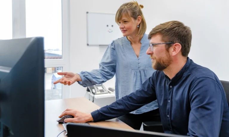 Eine Frau steht neben einem am Schreibtisch sitzenden Mann; beide blicken auf einen Computerbildschirm. Die Frau zeigt auf den Bildschirm, während sie über die Fallzahlberechnung sprechen. Im Hintergrund sind ein Bürotelefon und ein Whiteboard zu sehen.