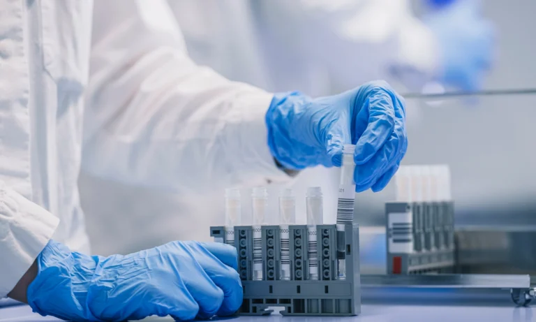 Eine Person, die blaue Handschuhe und einen weißen Laborkittel trägt, stellt ein Reagenzglas in ein Gestell, das bereits mit anderen Reagenzgläsern gefüllt ist, in einem Labor für Begleitdiagnostik.