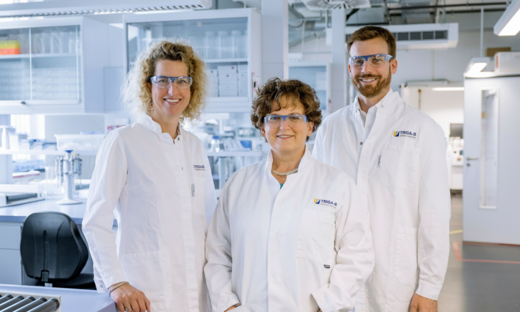 Drei Wissenschaftler in weißen Laborkitteln und Schutzbrillen stehen lächelnd in einem hellen, modernen Labor, im Hintergrund sind Laborgeräte und Regale zu sehen.