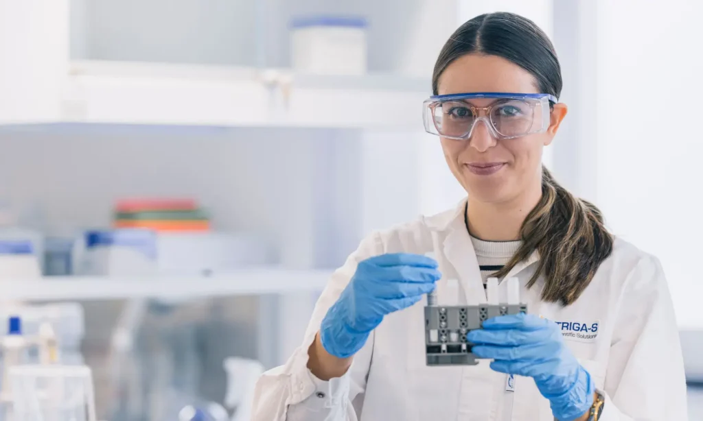 Ein Wissenschaftler mit Schutzbrille, blauen Handschuhen und weißem Laborkittel hält ein Probenfach in der Hand und lächelt in einem hell erleuchteten Labor, das für Spitzenleistungen im Bereich der Pharmadienstleistungen steht.