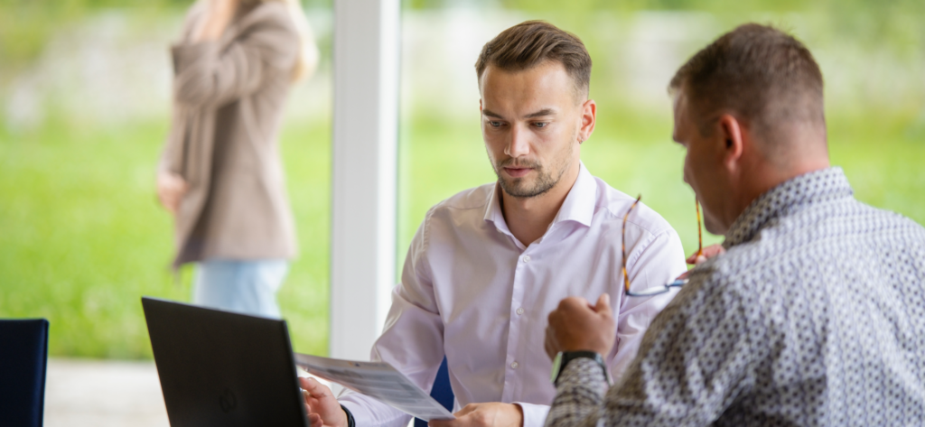Zwei Männer sitzen an einem Tisch und arbeiten gemeinsam an dmids; einer schaut auf einen Laptop, während der andere eine Brille und ein Dokument in der Hand hält. Im Hintergrund steht eine verschwommene Person in der Nähe eines großen Fensters, hinter dem sich Grünflächen erstrecken.