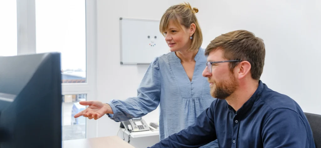 Eine Frau steht neben einem am Schreibtisch sitzenden Mann; beide blicken auf einen Computerbildschirm. Die Frau zeigt auf den Bildschirm, und es sieht so aus, als würden sie über Fallzahlberechnung sprechen. Im Hintergrund ist eine Whiteboard-Tafel zu sehen.