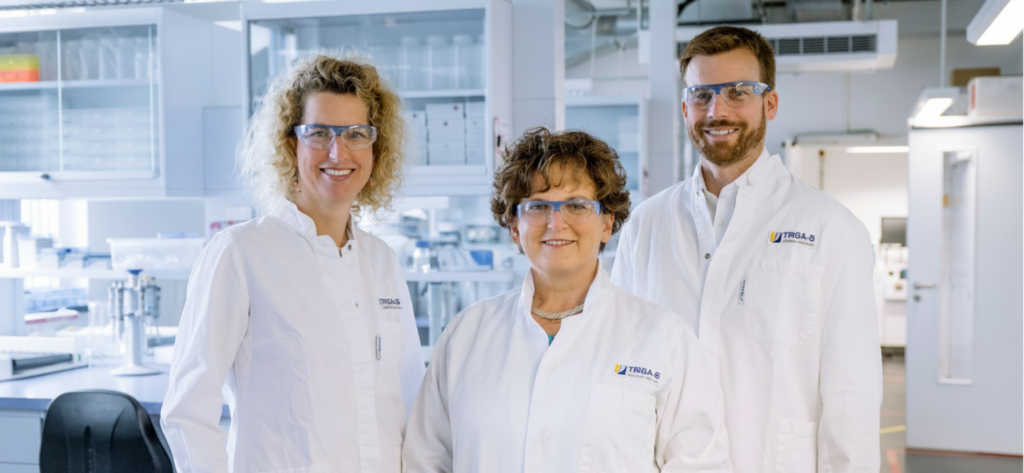 Drei Wissenschaftler in weißen Laborkitteln und Schutzbrillen stehen lächelnd in einem modernen Labor, umgeben von Laborgeräten und Regalen – eine Szene, die aus einem „Cro-kurz-erklärt“-Lehrvideo stammen könnte.