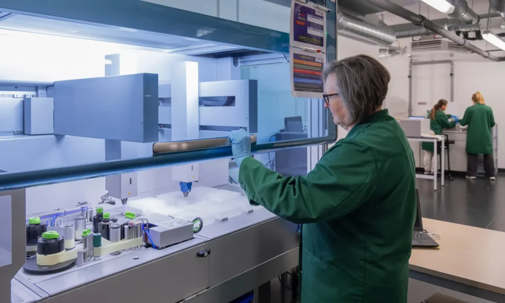 Ein Wissenschaftler in einem grünen Laborkittel bedient hochmoderne Laborgeräte, während zwei weitere im Hintergrund arbeiten – alles in einer modernen Cro-Wiki-Laborumgebung.