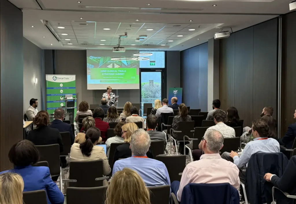 Während des „Clinical Trials Strategic Summit“ 2024 sitzt eine Gruppe von Menschen mit Blick auf einen Redner vorne im Konferenzraum, während auf einer Leinwand eine Präsentation gezeigt wird und an den Seiten Banner zu sehen sind.