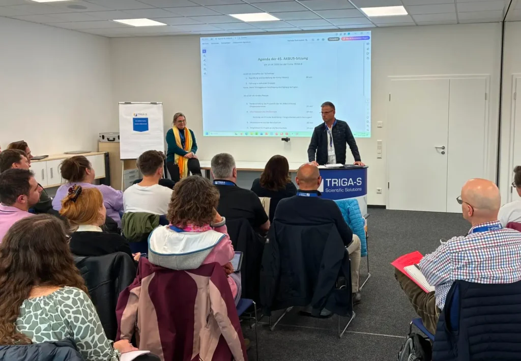 Ein Mann steht an einem Rednerpult und hält vor einem sitzenden Publikum in einem Konferenzraum beim „TRIGA-S auf dem LFAction!“-Symposium & Workshop einen Vortrag, während eine Frau daneben steht. Auf einer großen Leinwand wird die Tagesordnung angezeigt, und an der Seite steht ein Flipchart.