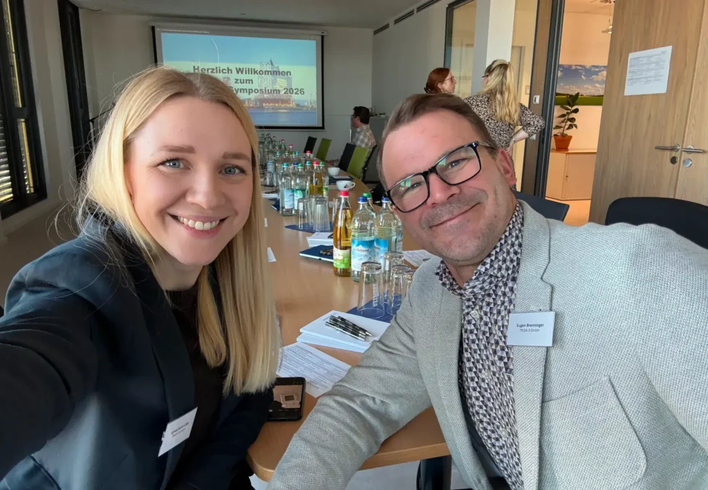 Zwei Personen in Geschäftskleidung lächeln für ein Selfie an einem Konferenztisch, auf dem Getränke und Unterlagen liegen. Hinter ihnen heißt eine Leinwand die Teilnehmer des „TRIGA-S auf dem LFAction!“-Symposiums und -Workshops willkommen; im Hintergrund sind weitere Teilnehmer zu sehen.