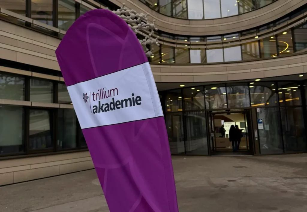 Vor einem modernen Gebäude mit großen Glasfenstern und geschwungener Architektur weht während des „TRIGA-S auf dem LFAction!“-Symposiums und -Workshops eine violette Fahne mit der Aufschrift „Trillium Akademie“; am Eingang sind Menschen zu sehen.
