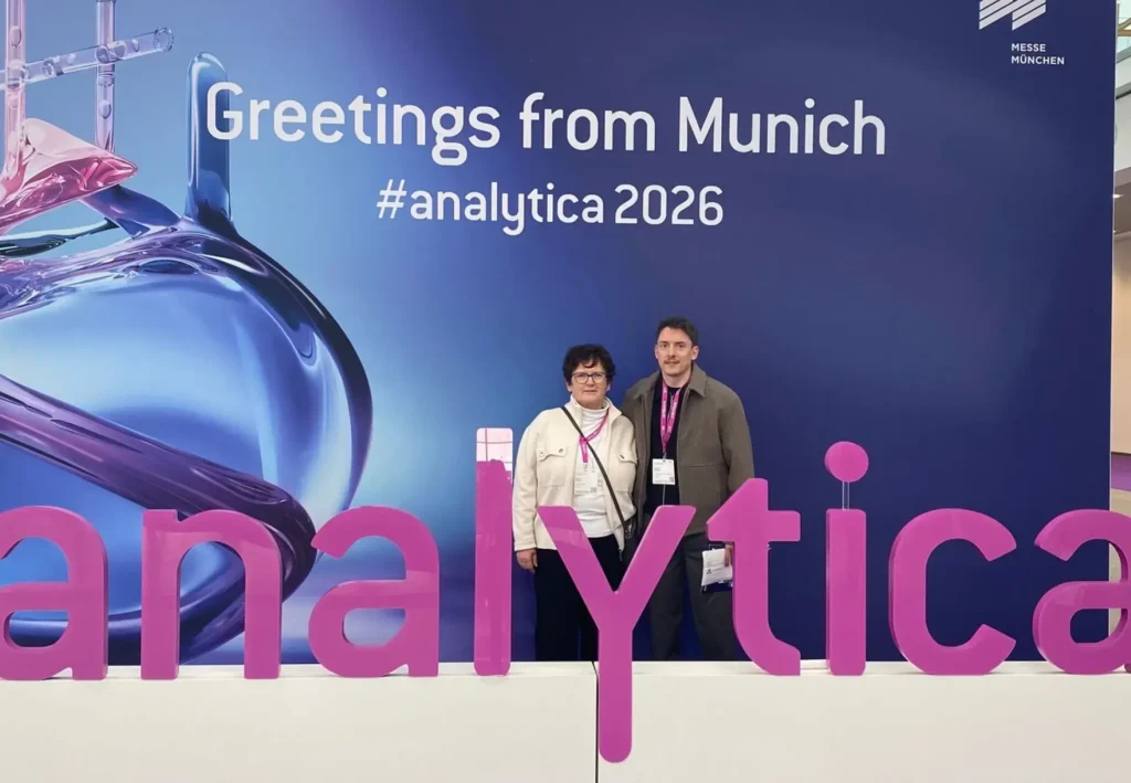 Zwei Personen stehen vor einem großen Schild mit der Aufschrift „Greetings from Munich #analytica2026“ beim TRIGA-S auf dem LFAction! Symposium & Workshop. Im Vordergrund ist ein Teil des Wortes „analytica“ in großen rosa Buchstaben zu sehen.