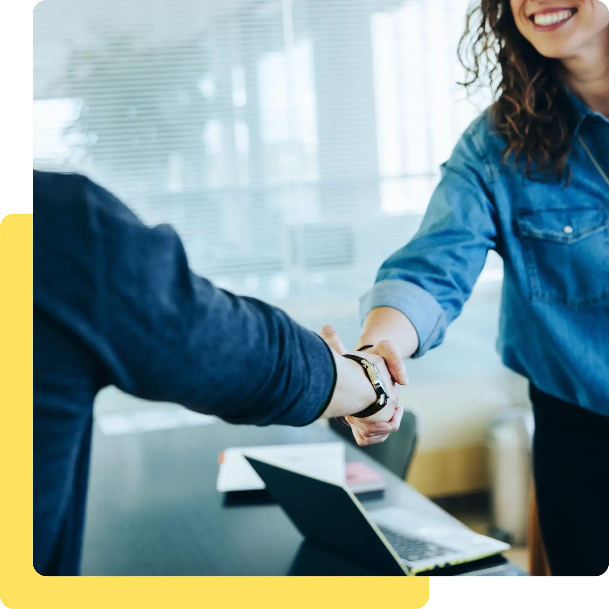 Two people shaking hands across a desk in a bright office, with a laptop in the foreground. Smiling, they celebrate a successful meeting—showcasing the collaborative spirit fostered by triga-s careers.