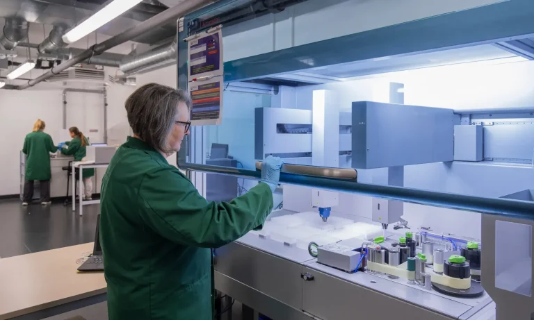 Ein Wissenschaftler in einem grünen Laborkittel und mit Handschuhen führt unter einem Abzug mit modernster Laborausstattung Leistungsstudien durch, während zwei weitere Forscher im Hintergrund eines modernen Labors arbeiten.