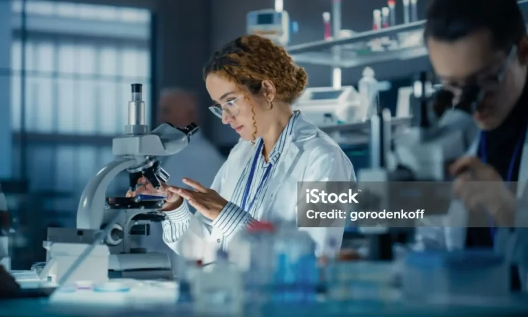 Eine Wissenschaftlerin in Laborkittel und Schutzbrille untersucht in einem modernen Auftragsforschungslabor eine Probe unter dem Mikroskop, während im Hintergrund ein weiterer Wissenschaftler arbeitet.