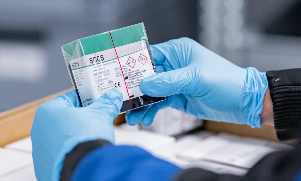 Eine Person mit blauen Handschuhen hält eine versiegelte Verpackung mit einer chemischen Probe in der Hand, die mit Gefahrenzeichen und Etiketten zur EU-Konformitätsbewertung versehen ist, und untersucht diese sorgfältig in einer Umgebung, die wie ein Labor oder ein Lagerraum aussieht.