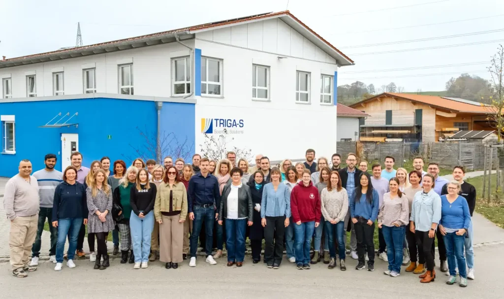Eine große Gruppe von Menschen posiert gemeinsam vor einem weiß-blauen Gebäude mit der Aufschrift „TRIGA-S“ und lächelt an einem sonnigen Tag in die Kamera. Das Treffen unterstreicht ihre Zusammenarbeit im Bereich der Auftragsforschung, bei der innovative Köpfe für Spitzenforschung im Auftrag zusammenkommen.