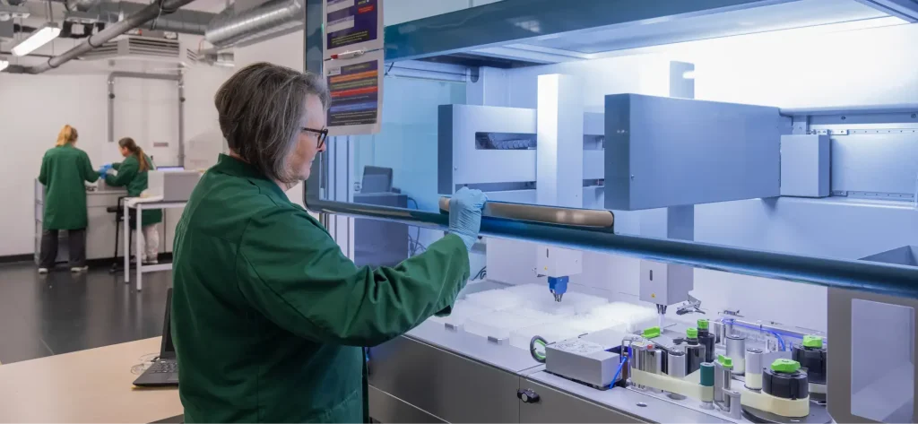 Ein Wissenschaftler in einem grünen Laborkittel führt Leistungsstudien mit automatisierten Laborgeräten durch, während zwei weitere Personen im Hintergrund eines modernen Labors arbeiten, das mit wissenschaftlichen Instrumenten und Verbrauchsmaterialien ausgestattet ist.