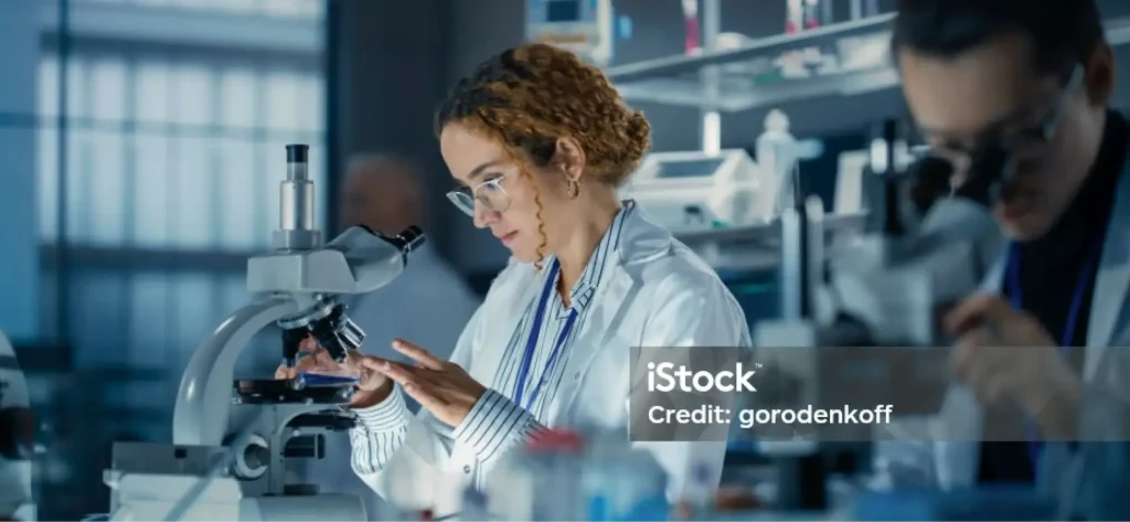 Ein Wissenschaftler mit lockigem Haar und Brille untersucht in einem Auftragsforschungslabor eine Probe unter dem Mikroskop. Im Hintergrund arbeitet ein weiterer Forscher, umgeben von Laborgeräten und Regalen.
