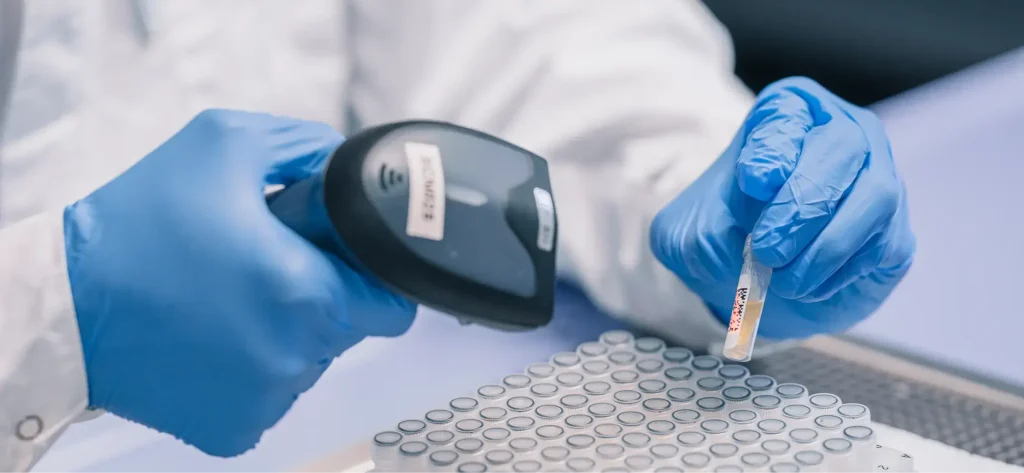 Eine Person mit blauen Handschuhen scannt einen Barcode auf einem kleinen Reagenzglas und hält es dabei über ein Tablett, das mit ähnlichen Reagenzgläsern gefüllt ist, um in einem Labor die bestimmungsgemäße Verwendung jeder Probe zu überprüfen.