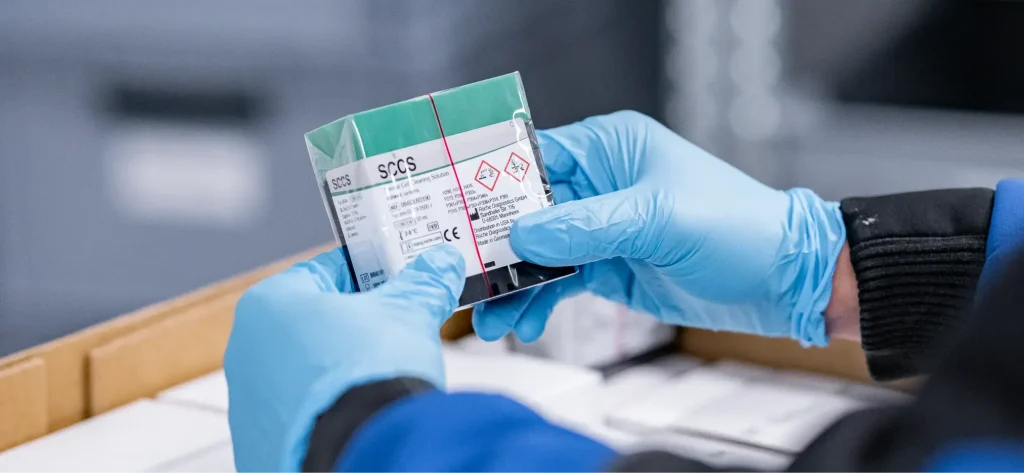 Mit Handschuhen bekleidete Hände halten ein kleines, versiegeltes Päckchen, das mit Chemikaliengefahrensymbolen und einem EU-Konformitätszeichen gekennzeichnet ist, in einer Lager- oder Laborumgebung, im Hintergrund sind Kartons zu sehen.